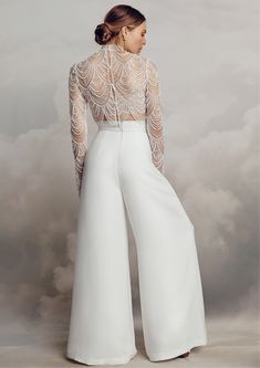 a woman wearing white pants and a sheer top with an open back, standing in front of clouds