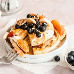 a white plate topped with pancakes covered in blueberries and peaches next to a cup of coffee