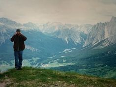 a man standing on top of a lush green hillside next to mountains covered in snow