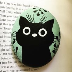 a rock with a black cat painted on it's face and eyes, sitting in front of an open book