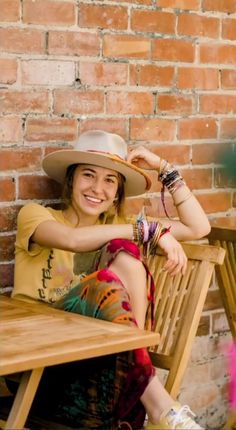 a woman wearing a hat sitting on a wooden bench next to a brick wall smiling