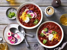 two bowls of chili with cheese, onions and other toppings on a table next to drinks