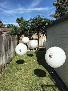 some white balloons are in the grass near a house and fence with trees behind them