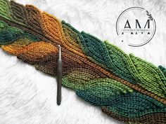 a crocheted feather is shown with a needle in the foreground and an am logo above it