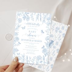 a person holding up two blue and white wedding cards with floral designs on the front