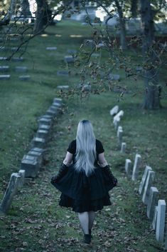 a woman with long white hair walking down a path in a cemetery surrounded by tombstones