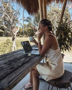 a woman sitting at a picnic table with a laptop and cup in her hand while looking into the distance