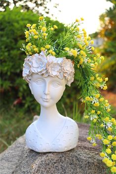 a white statue with yellow flowers on it's head sitting on top of a rock