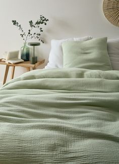 a bed with green sheets and pillows next to a wooden side table in a white room