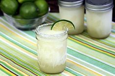 two margaritas sitting on top of a table next to some limes and jars