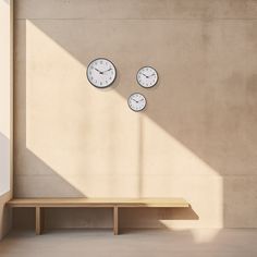 three clocks on the wall above a bench in an empty room with light coming through