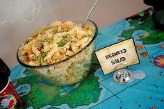 a glass bowl filled with pasta salad next to a bottle of wine and a sign