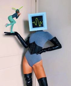 a woman in a bodysuit and boots with a monitor on her head, standing next to a wall