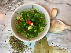 a white bowl filled with green vegetables next to garlic and an ear of garlic on a marble counter top