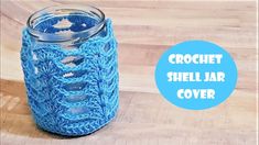 a blue crochet shell jar cover sitting on top of a wooden table next to a mason jar