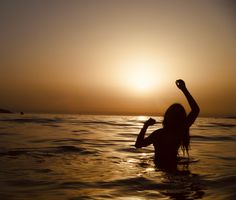 a woman standing in the ocean at sunset raising her arms up to the sky with one hand