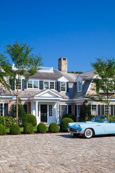 a classic car parked in front of a large house with hedges and trees on the driveway