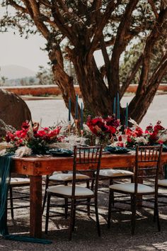 a wooden table topped with lots of red flowers and greenery next to a tree