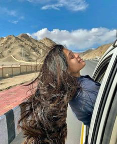 a woman leaning out the window of a car looking up at mountains in the distance