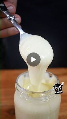 a person holding a spoon over a jar filled with liquid and cream on top of a wooden table