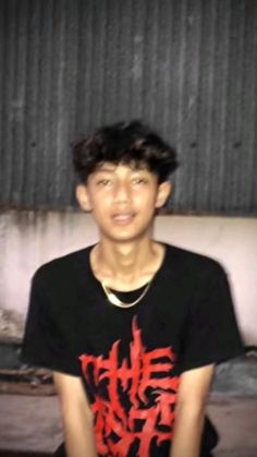 a young man sitting on top of a wooden floor next to a metal wall and wearing a black t - shirt