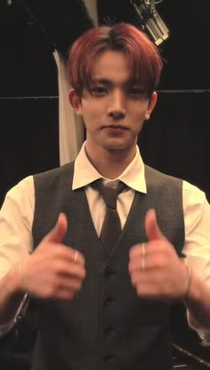 a man with red hair wearing a vest and tie giving the thumbs up sign in front of him