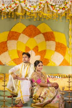 a man and woman sitting next to each other in front of a wall with flowers