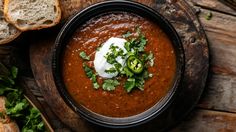 a bowl of red chili with sour cream and corian seeds on top, next to some bread