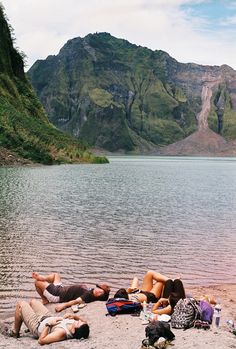 several people laying on the sand near a body of water with mountains in the background