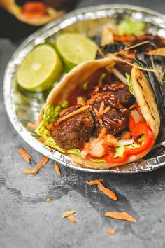 two tacos with meat, vegetables and sauce on a metal plate next to lime wedges