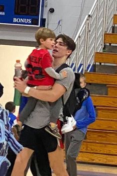 a man holding a young boy in his arms while standing on a basketball court with other people watching