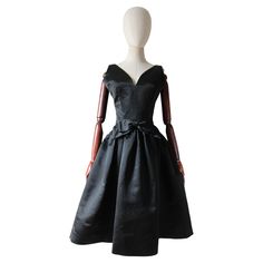 This truly mesmerising 1950's couture dress, rendered in a sumptuous black satin and accented by a decorative central bow, is just the piece to add to your timeless capsule collection. The elegant curved V shaped neckline is framed by medium shoulder straps and a sleeveless cut. The fitted bodice is accented by tailored darts along the bust-line, and structural seams that run upwards from the dropped skirt seam. The cinched in waistline, sets off the lowered skirt seam, and large central black satin bow. The elegant line of the skirt is set off by box pleats that drape from either side of the central box, completing this dress in a truly iconic 1950's silhouette. The back of the dress is accented by a deep V backline, framed to either side by tailored seams. This dress is lined in silk org 1940s Evening Dresses, Lame Dress, Evening Dresses Uk, Satin Noir, Satin Evening Dresses, Cocktail Dress Vintage, Duchess Satin, Red Velvet Dress, Couture Dress