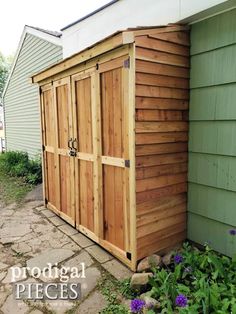 a wooden storage shed sitting next to a green house with purple flowers in the front yard