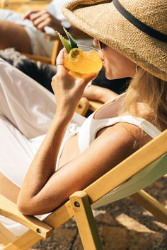 a woman sitting in a lawn chair drinking from a wine glass while wearing a straw hat