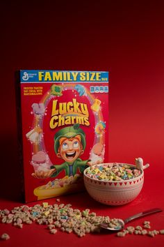 lucky charms cereal box next to bowl of cereal and spoon on red background with family size book