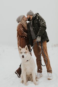 a man and woman kissing in the snow with a husky dog on a leash next to them