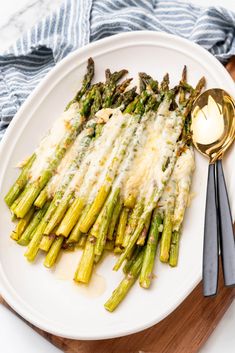 asparagus covered in melted cheese on a white plate with silver spoons next to it