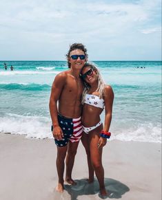 two people are standing on the beach in swimsuits and one is wearing sunglasses