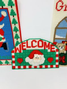 three cross stitch christmas welcome signs with santa clause and snowman on the front, one in red white and green