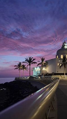 the building is white and has palm trees in front of it at dusk with purple clouds