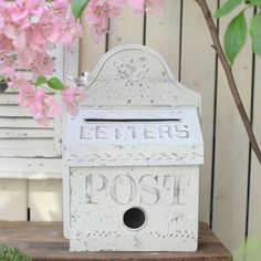 a white mailbox sitting on top of a wooden bench next to pink flowers and a tree