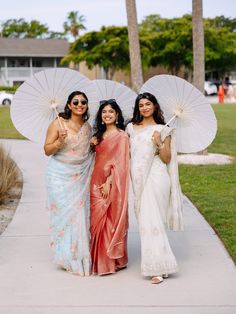 three women in sari holding umbrellas and posing for a photo on the sidewalk