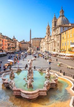 a fountain in the middle of a city square with people walking around and buildings on either side