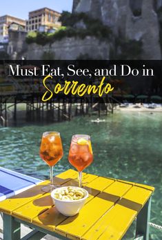 two glasses of wine sitting on top of a yellow table next to the ocean with text overlay that reads must eat, see and do in sorrento