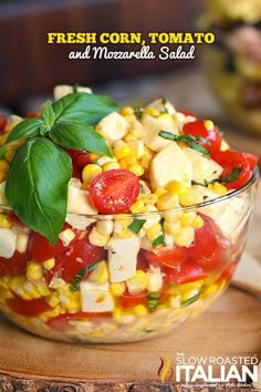 a salad with tomatoes, corn and basil in a glass bowl on a cutting board