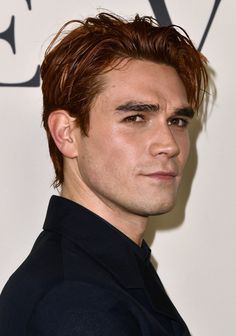 a close up of a person wearing a black suit and tie with red hair in front of a white wall