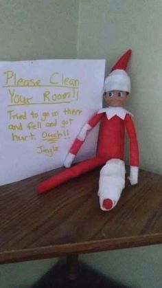 an elf is sitting on a table with a sign in front of him that says please clean your room