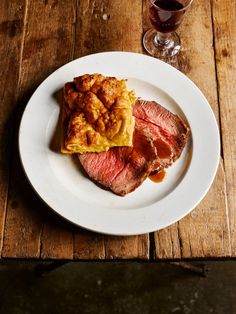 a white plate topped with meat and potatoes next to a glass of wine on top of a wooden table