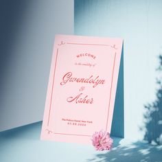 a pink wedding welcome card sitting on top of a blue table next to a tree