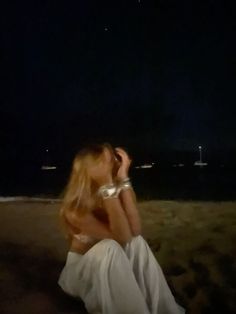 a woman is sitting on the beach at night with her hands in her hair and looking off into the distance